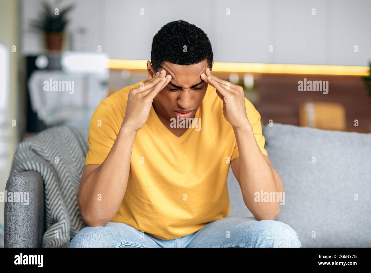 Disappointment latino guy in a yellow t-shirt sits on a sofa at home, massages his temples with his eyes closed, he has personal problems, headache, migraine, stress, thinks about solving the problem Stock Photo