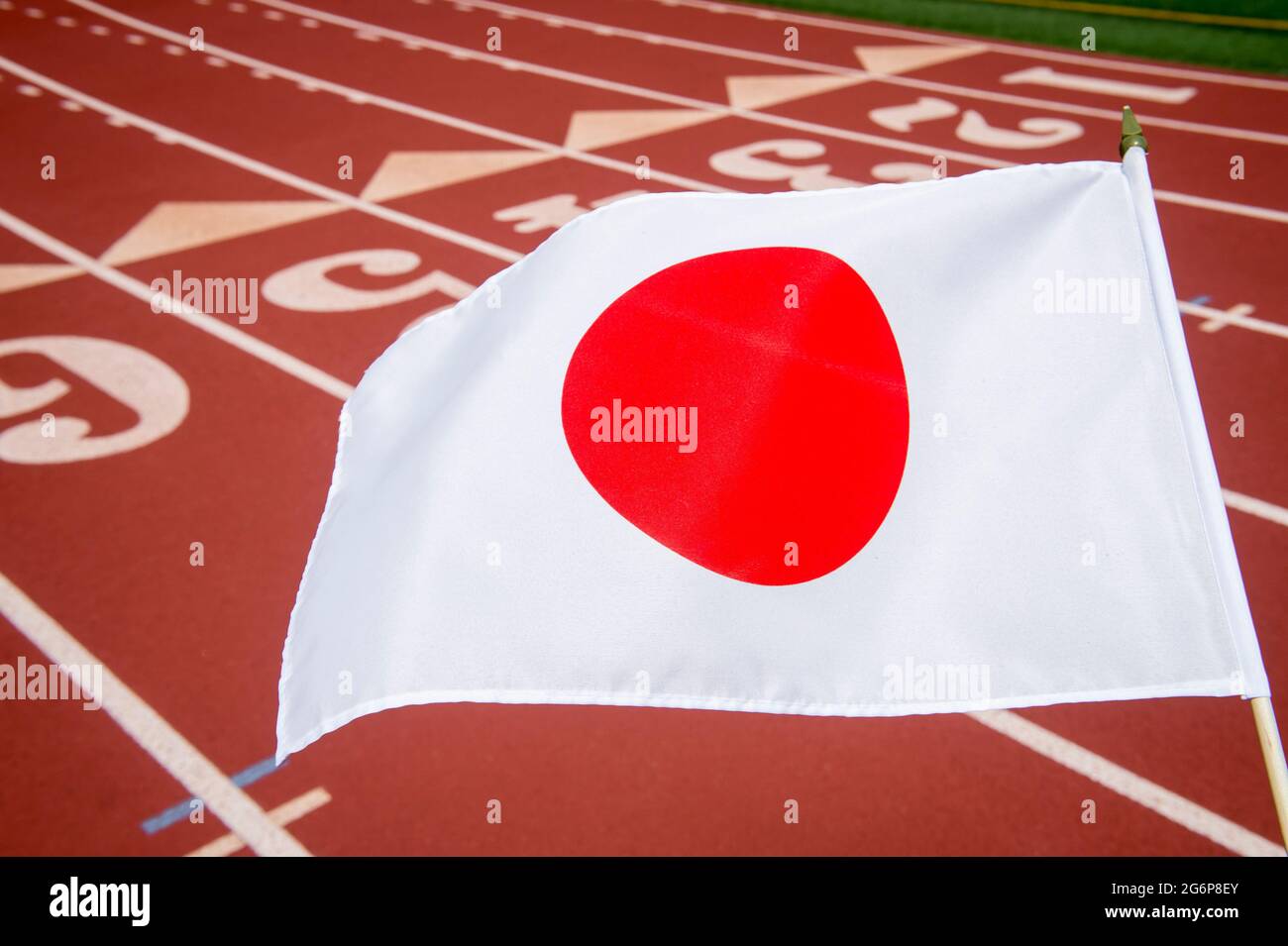 Bright sunny view of a Japanese flag flying in front of the numbered lanes at the starting line of a red running track Stock Photo