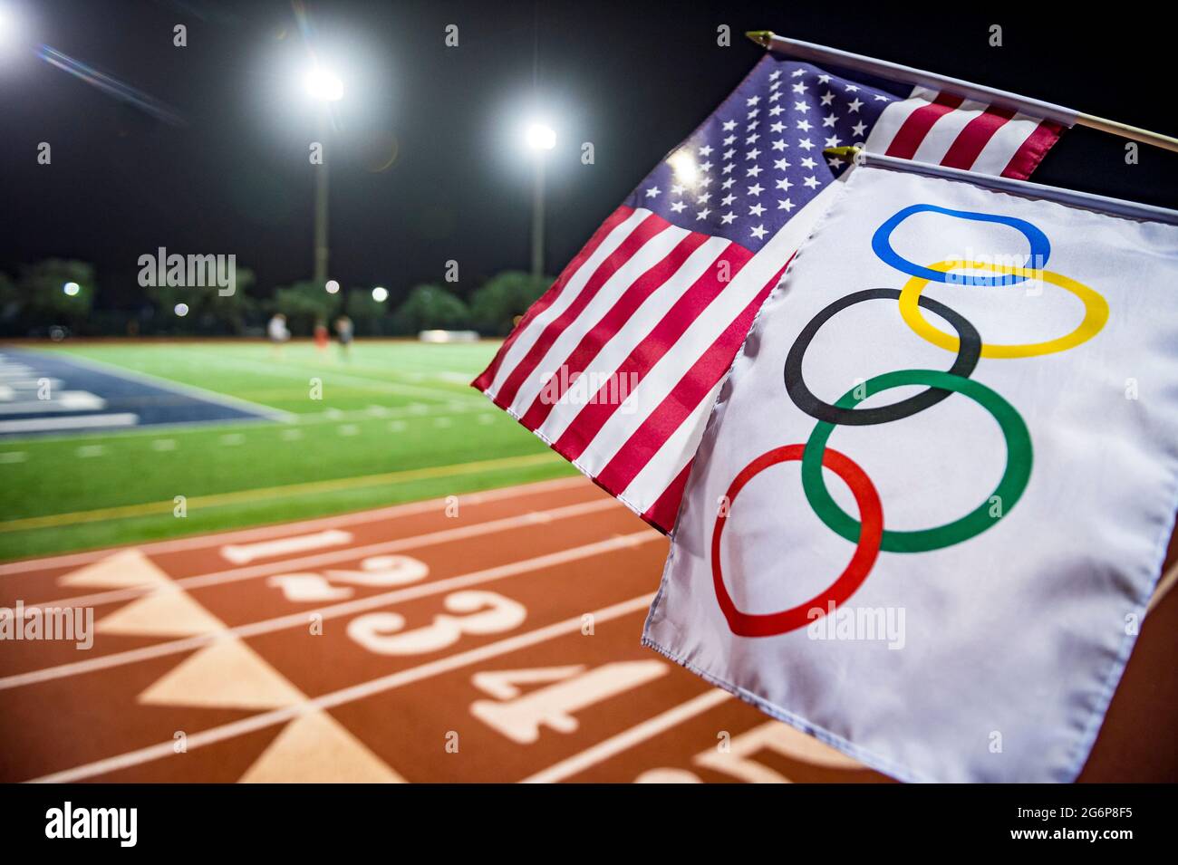 MIAMI, USA - AUGUST 15, 2019: An Olympic and American flag wave together under the floodlights of a red athletics track. Stock Photo