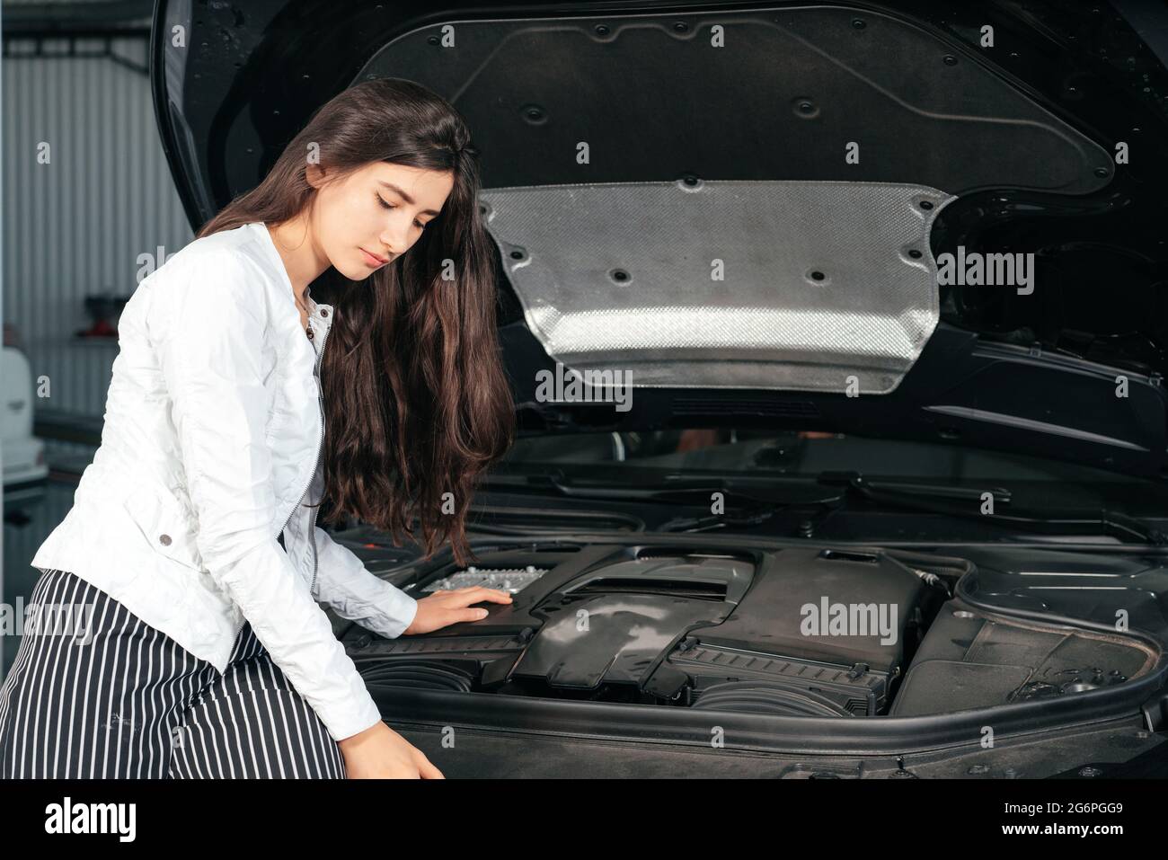 Young woman standing in front of the car with opened hood in garage
