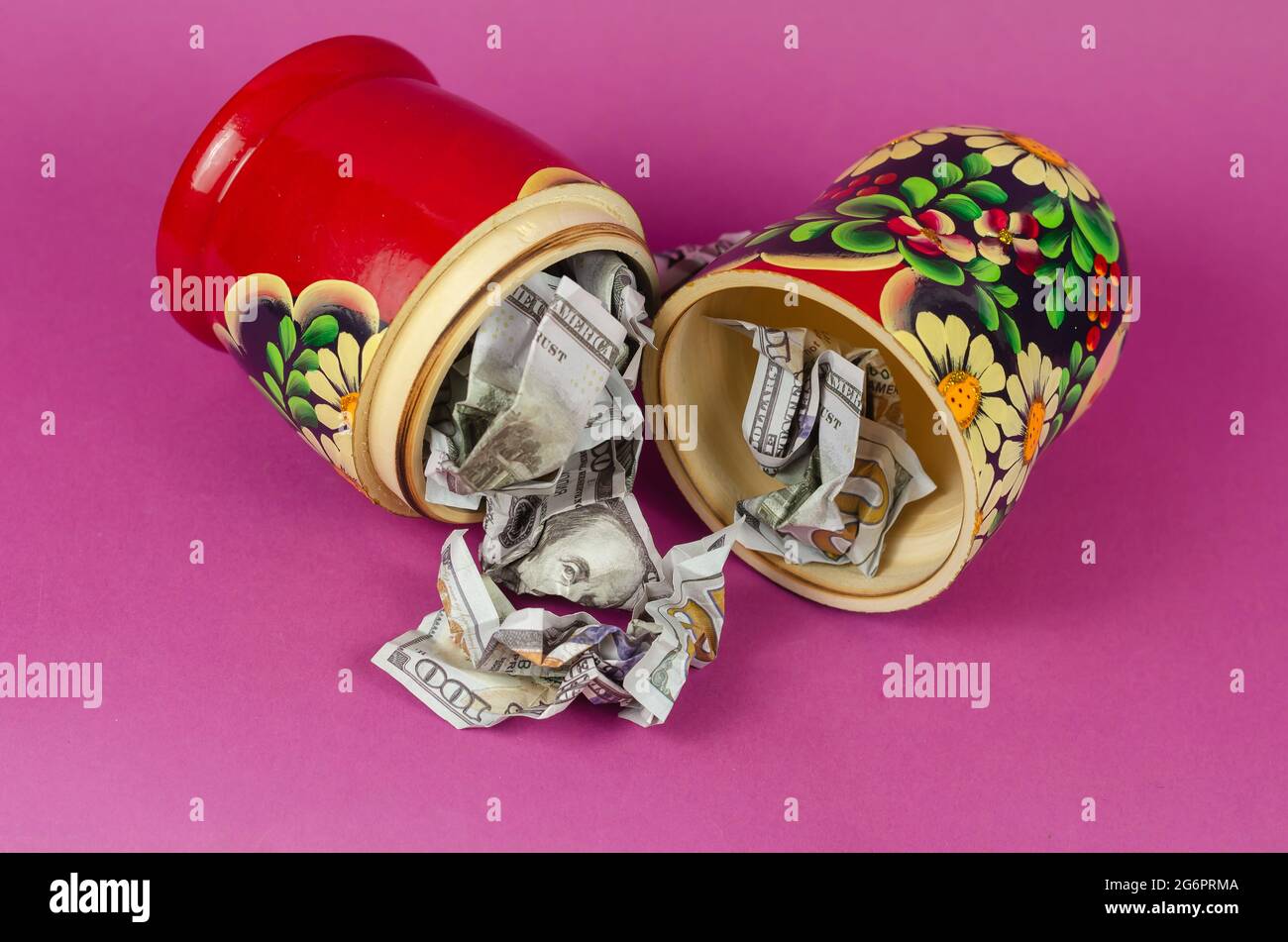 xRussian matryoshka full of money. Crumpled hundred-dollar bills fall out of open wooden toy. Symbol of Russia and American money on  pink background Stock Photo
