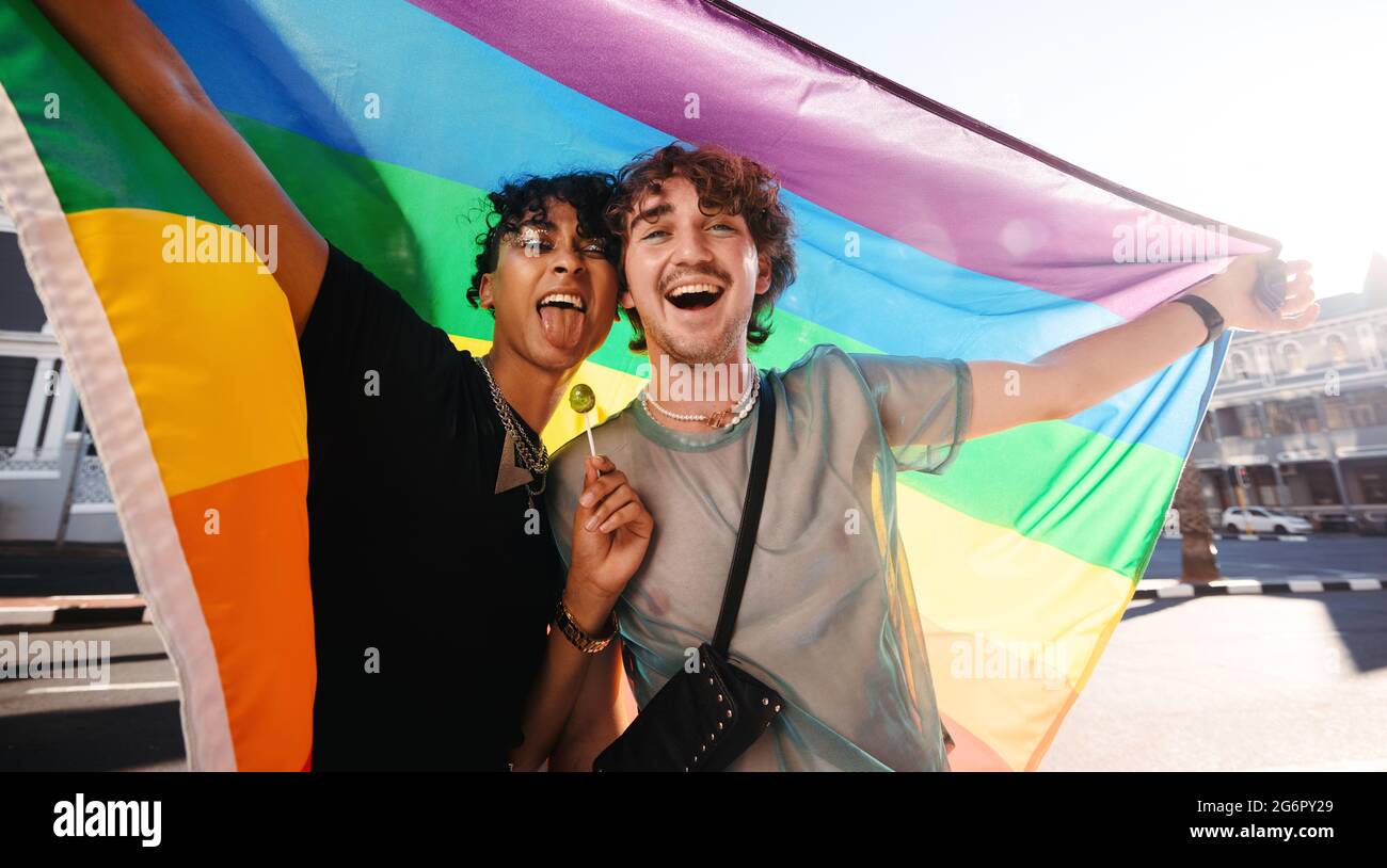 Non-conforming young men celebrating gay pride. Two young gay men smiling cheerfully while raising a rainbow pride flag. Two members of the LGBTQ+ com Stock Photo