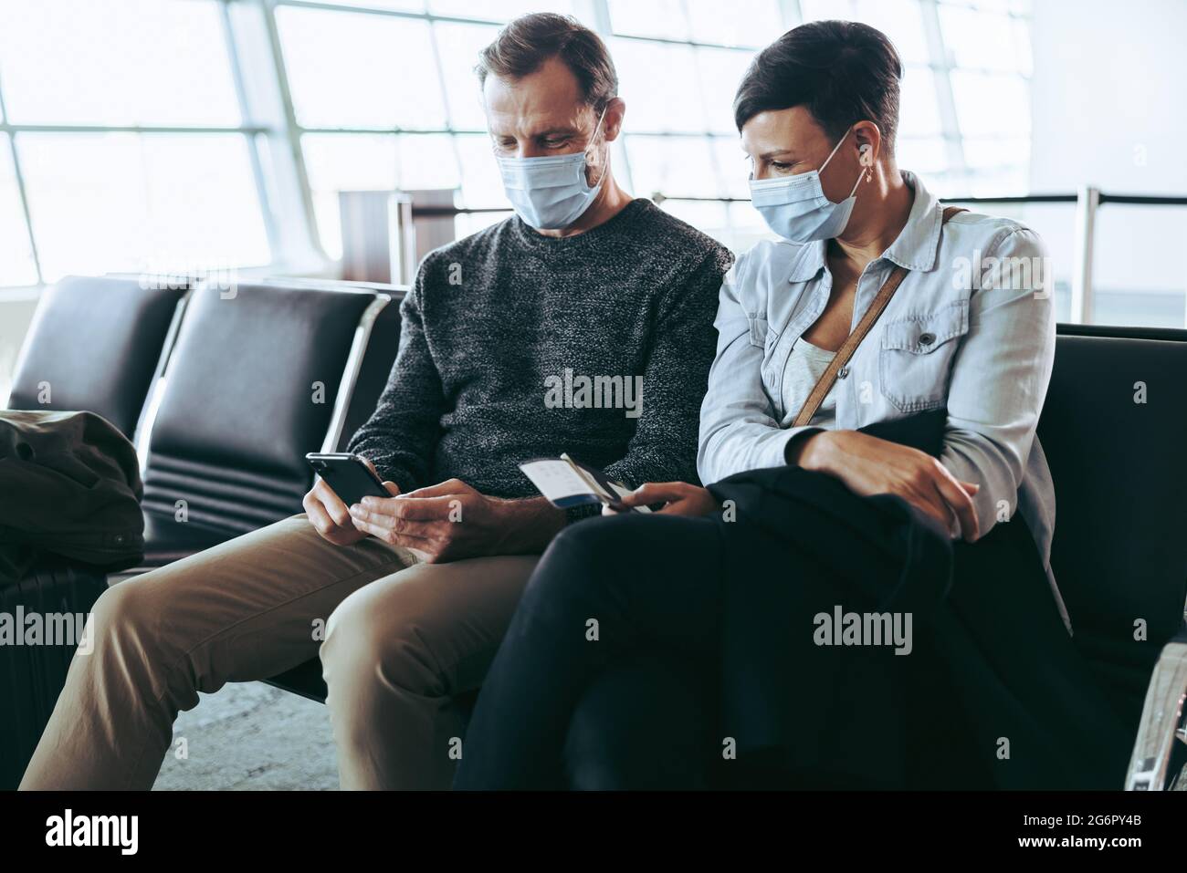 Couple wearing face masks checking mobile phone for flight timing while waiting at airport terminal. Man and woman at airport during corona virus pand Stock Photo