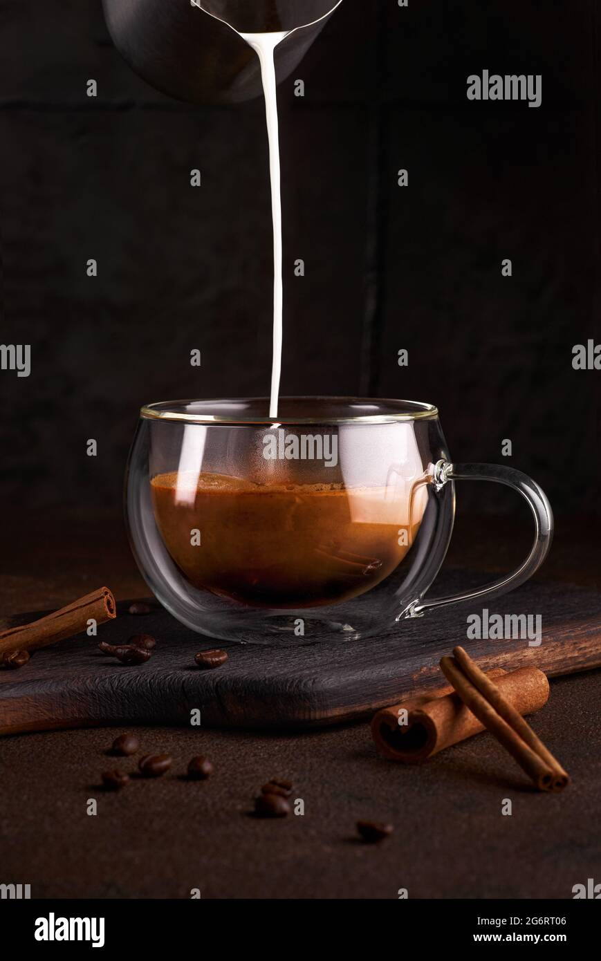 Coffee with milk in glass at dark background. Hand pouring milk cream