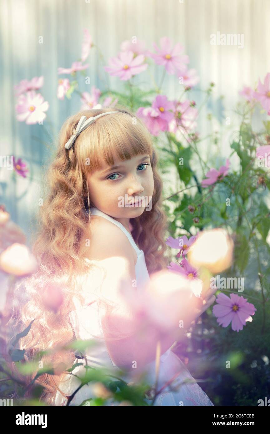 Portrait of a 10-year-old little girl in a blooming garden. A cute beautiful baby with beautiful long curly red hair is having fun with the flowers of Stock Photo