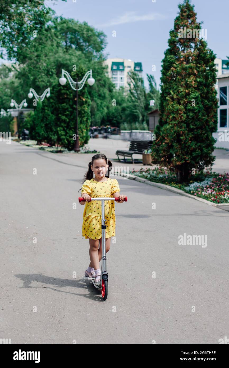 a cute girl in a yellow sundress learns to ride a scooter around the city and enjoys a hot summer day Stock Photo