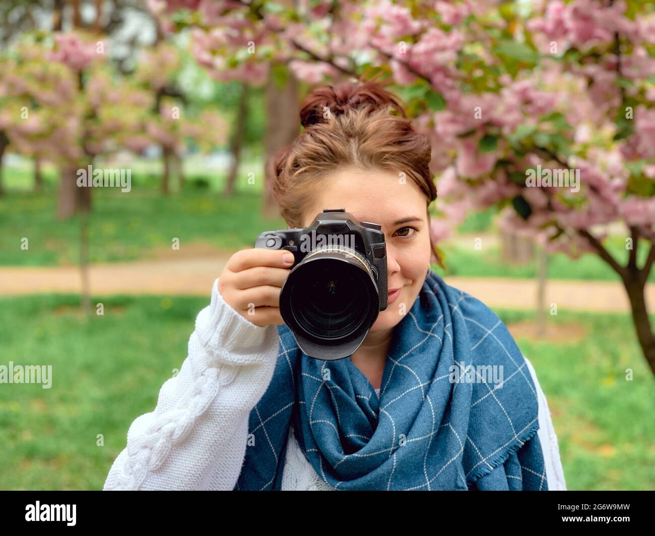 young happy smiling red-haired woman with a white sweater and blue scarf holds in her hands and looks at the camera near a blossoming pink sakura tree Stock Photo