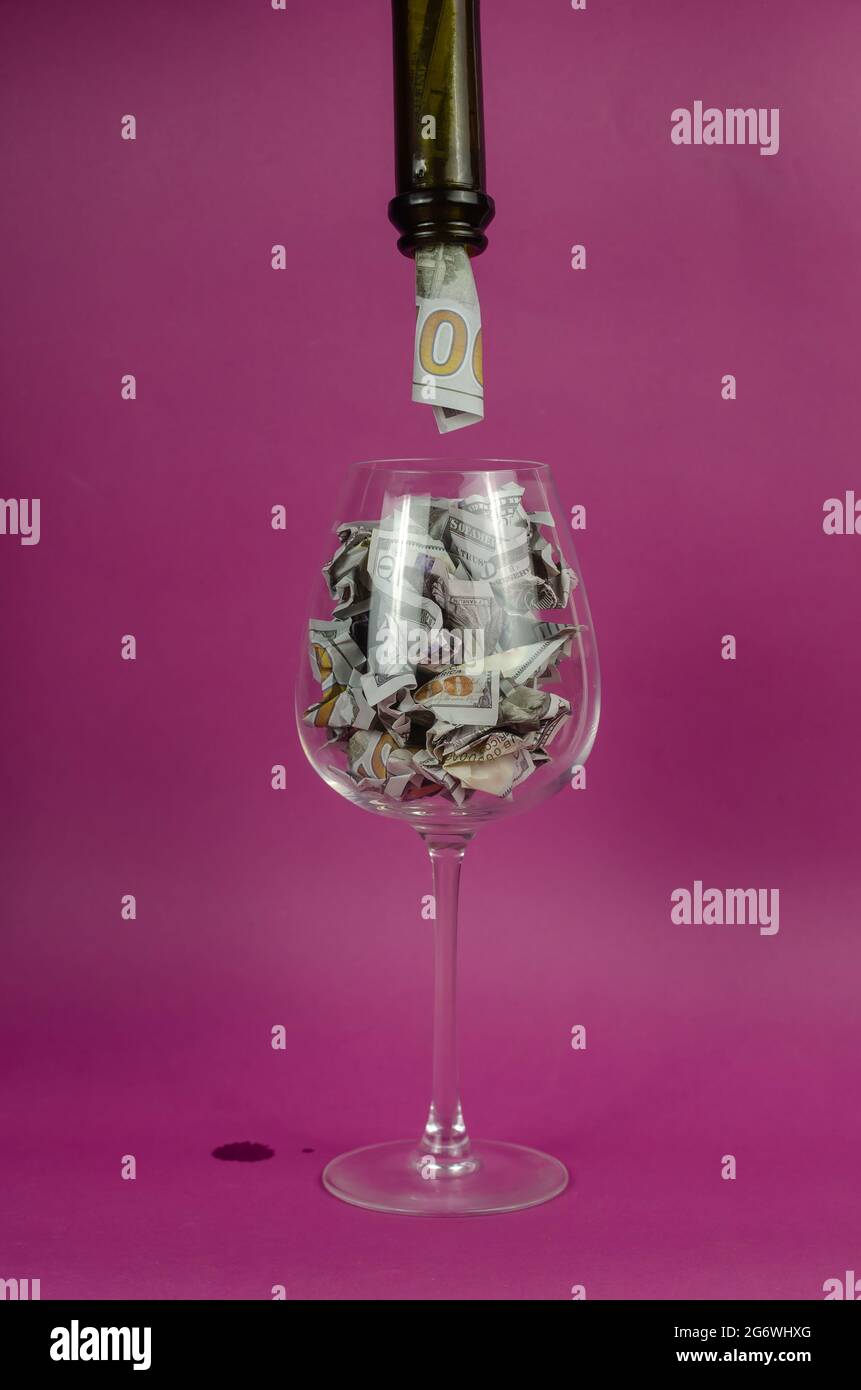 Wine glass is filled with money from bottle. WineGlass is full Crumpled hundred dollar bills on pink background. Stock Photo