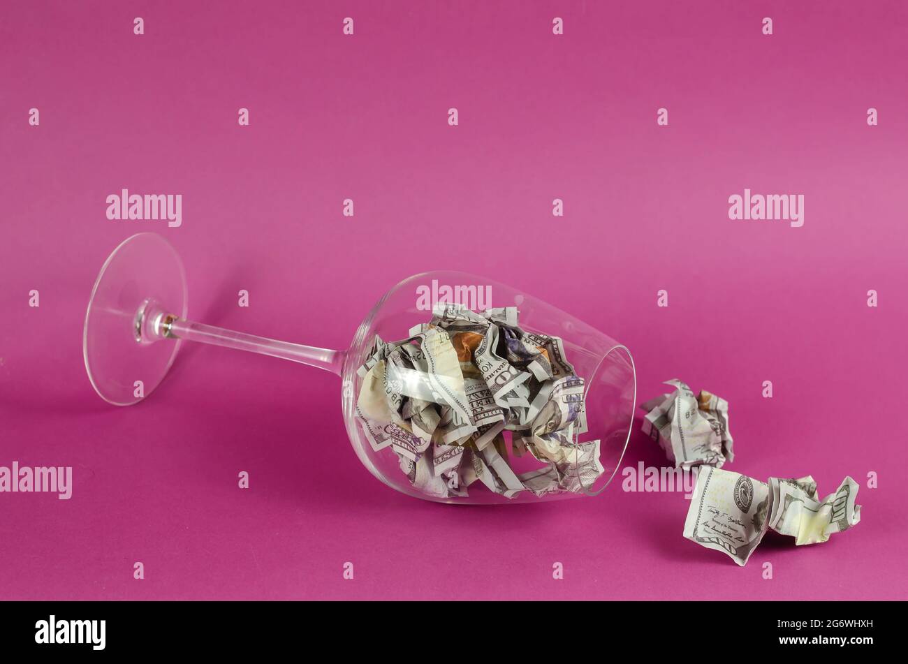 Transparent wine glass with money lying on pink background. Glass is full Crumpled hundred dollar bills. Stock Photo