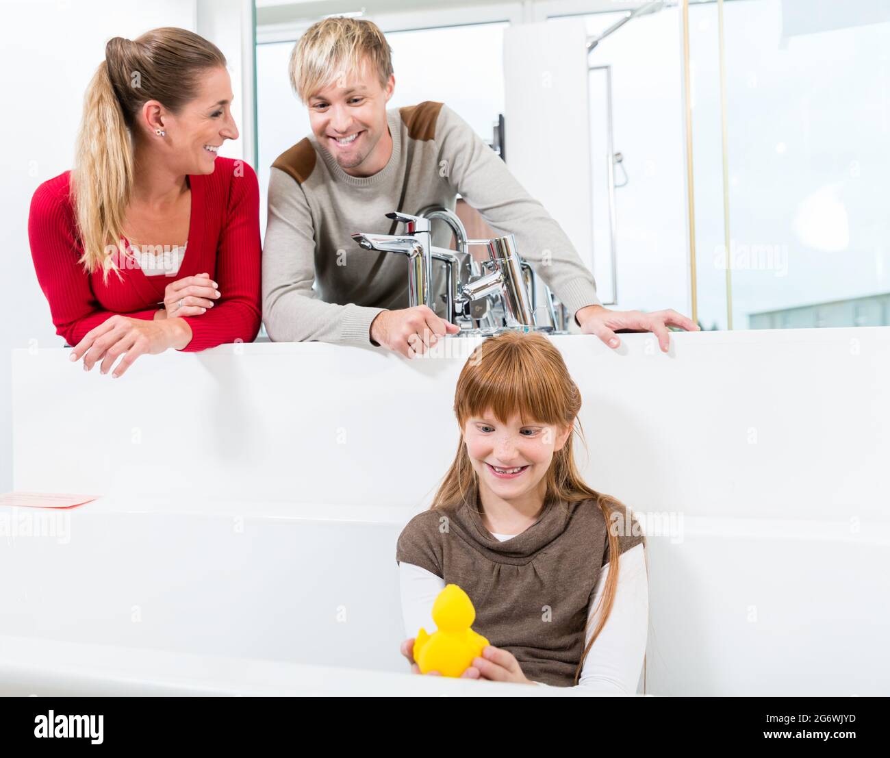 Cute and funny girl looking at her happy parents, while sitting in a white bathtub in the showroom of a sanitary ware shop with high-quality fixtures Stock Photo