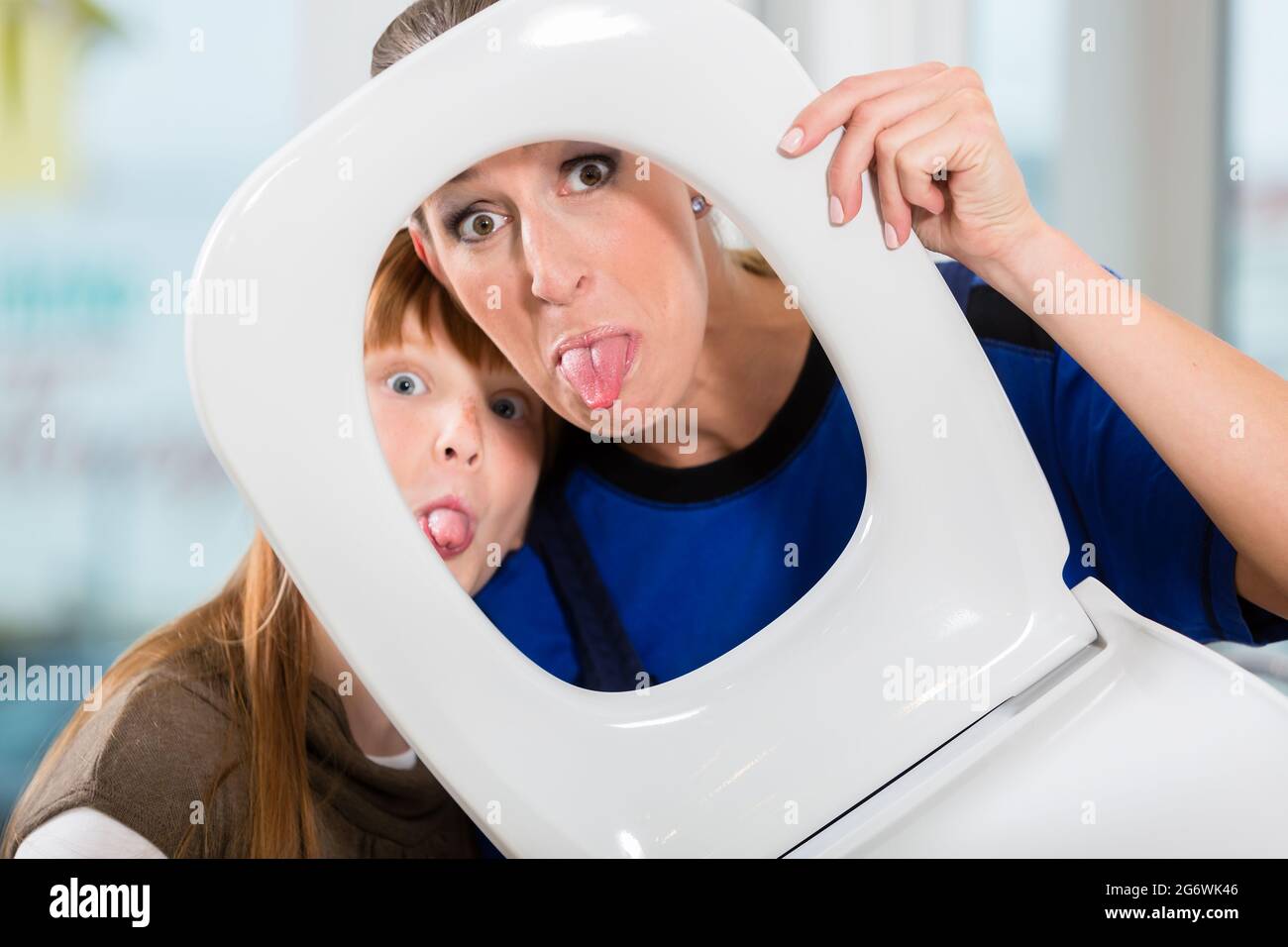 Funny portrait of a cheerful woman and her cute daughter smiling and looking at camera through a white toilet seat in a sanitary ware shop Stock Photo