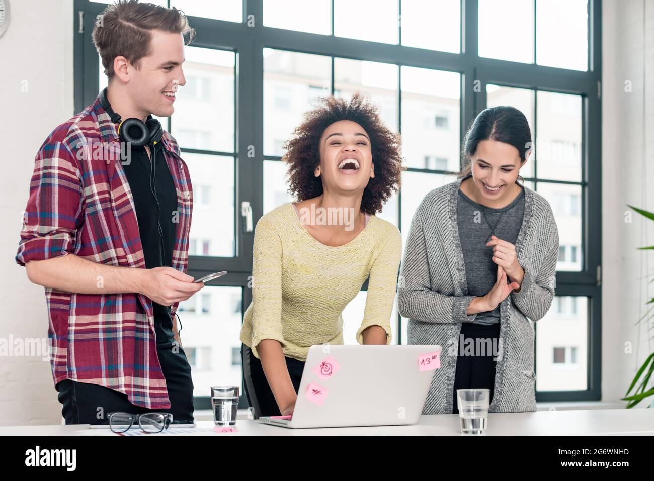 Three young colleagues laughing while standing in front of a laptop in the meeting room of a modern company Stock Photo