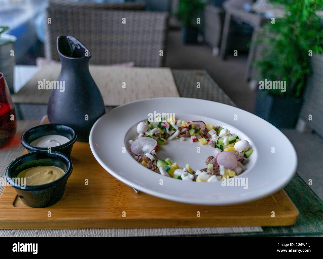 Ingredients for okroshka, a Russian cold soup, served on a white plate, with kvas jar standing beside to be added by a patron. Stock Photo