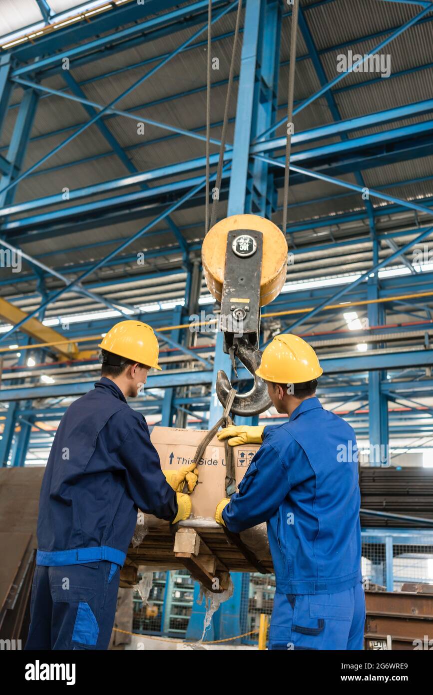 Two Asian workers handling heavy loading lifted by crane in the interior of a metallurgical factory Stock Photo