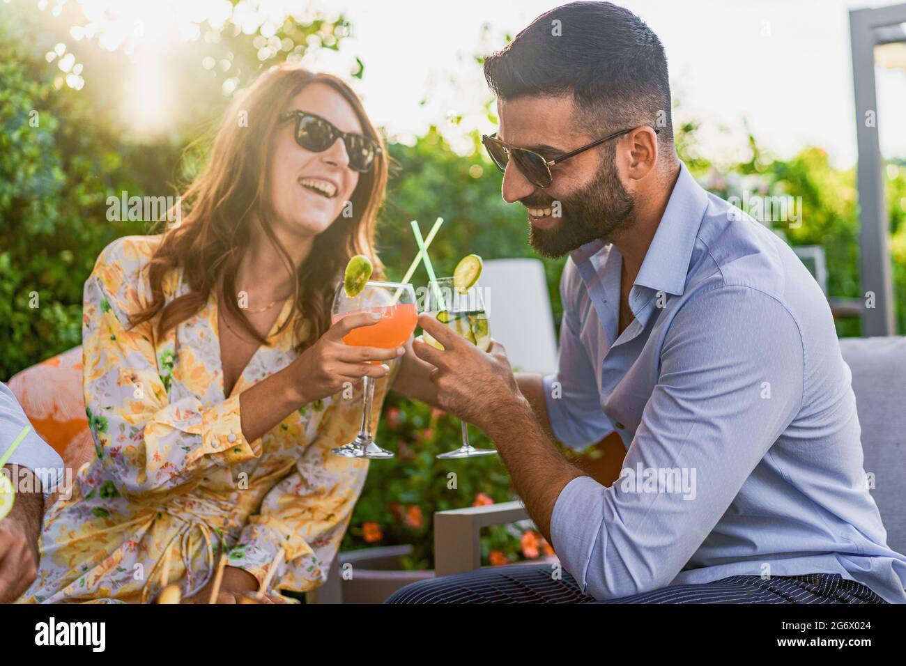Couple of young friends having a celebratory toast with clinking glasses with fruit cocktails while sitting on a sofa at a garden party. Young people Stock Photo