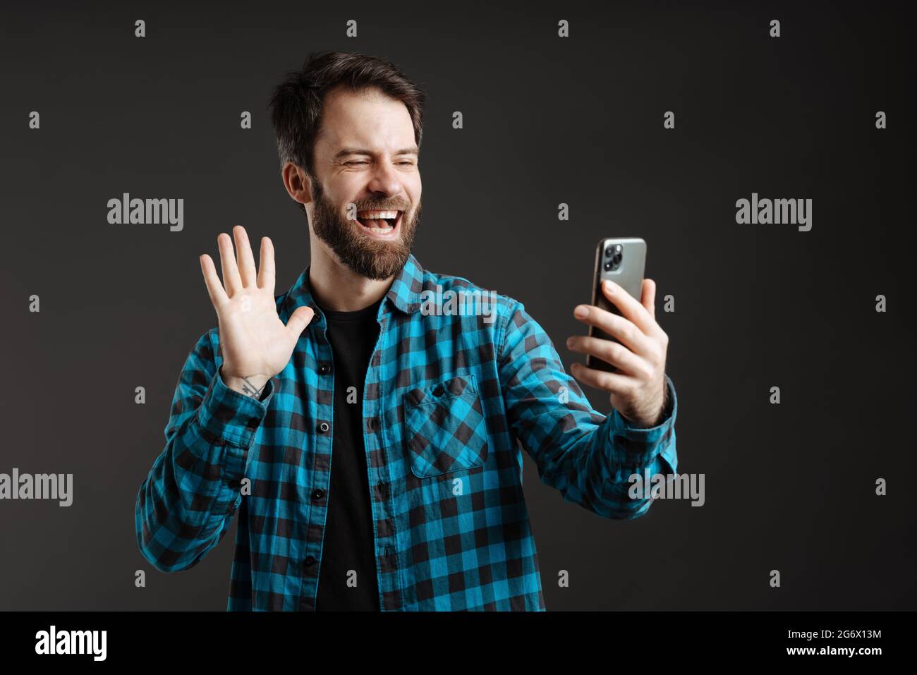 Bearded happy man waving hand while using mobile phone isolated over black background Stock Photo
