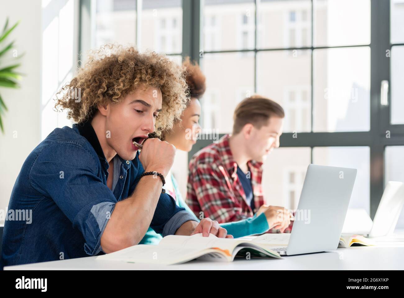 Side view portrait of a funny student yawning in front of a book, while sitting down at desk in the classroom at a modern college or university Stock Photo