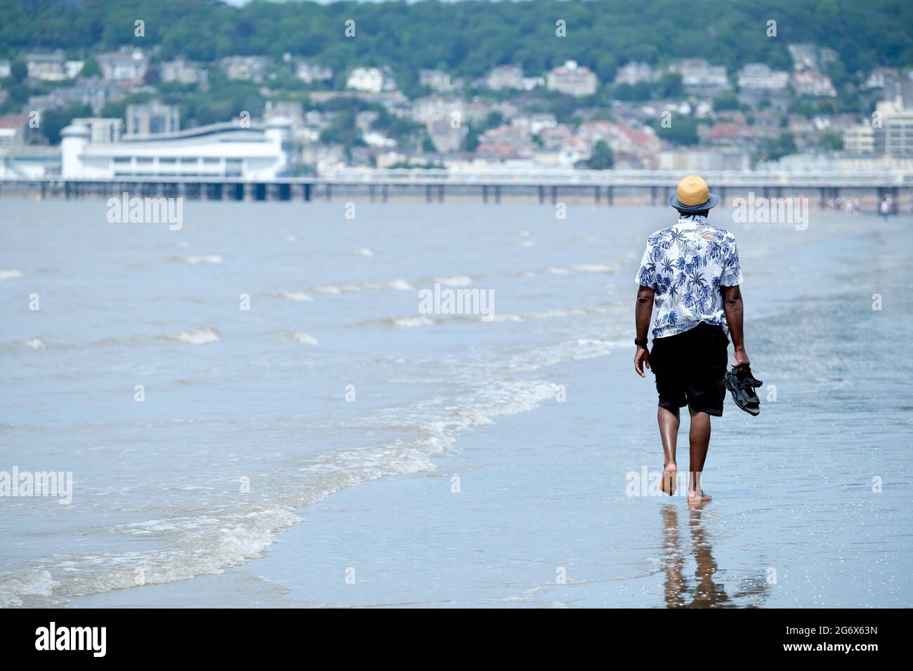 A male tourist paddles, barefooted, along the shoreline at Weston super mare beach UK. The towns pier can be made out in the background Stock Photo
