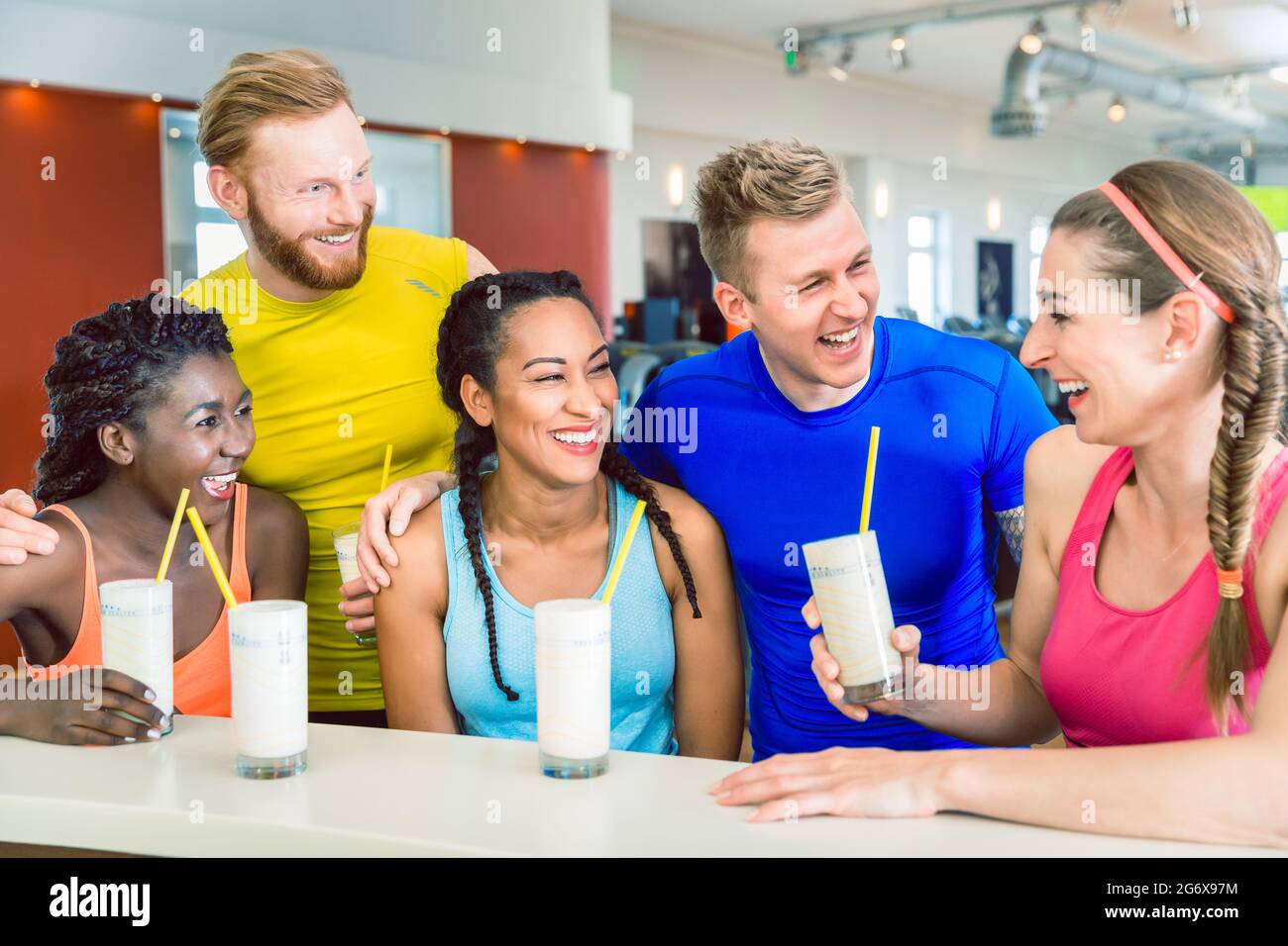 Multi-ethnic group of cheerful friends laughing while drinking white protein shakes before workout at a modern fitness club Stock Photo