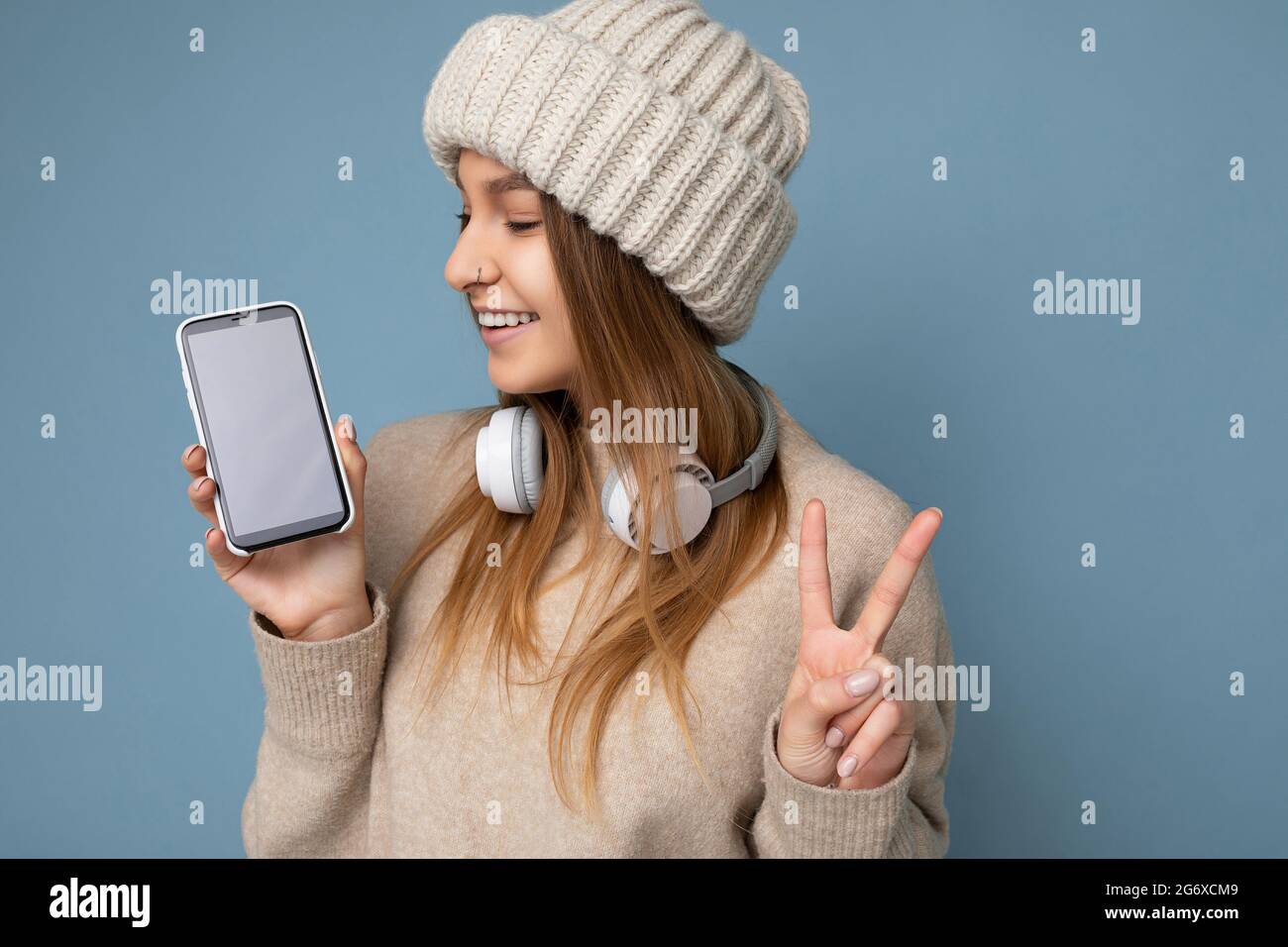 Closeup portrait Photo of beautiful positive joyful young dark blonde woman wearing beige stylish sweater and knitted hat wearing earphones and Stock Photo