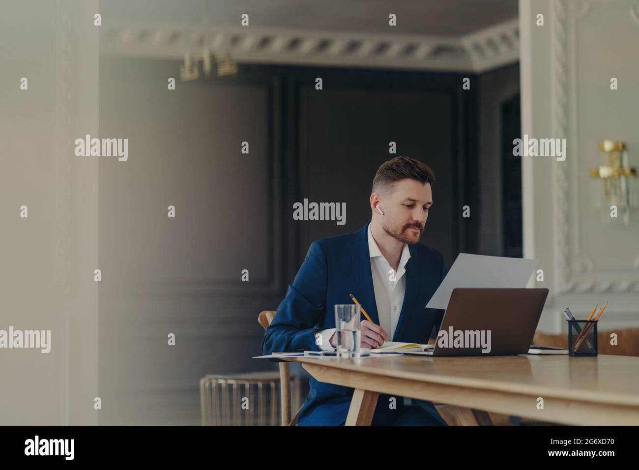 Serious businessman in smart suit writes in notepad concentrated at paper document poses at office desk with laptop computer glass of water uses Stock Photo