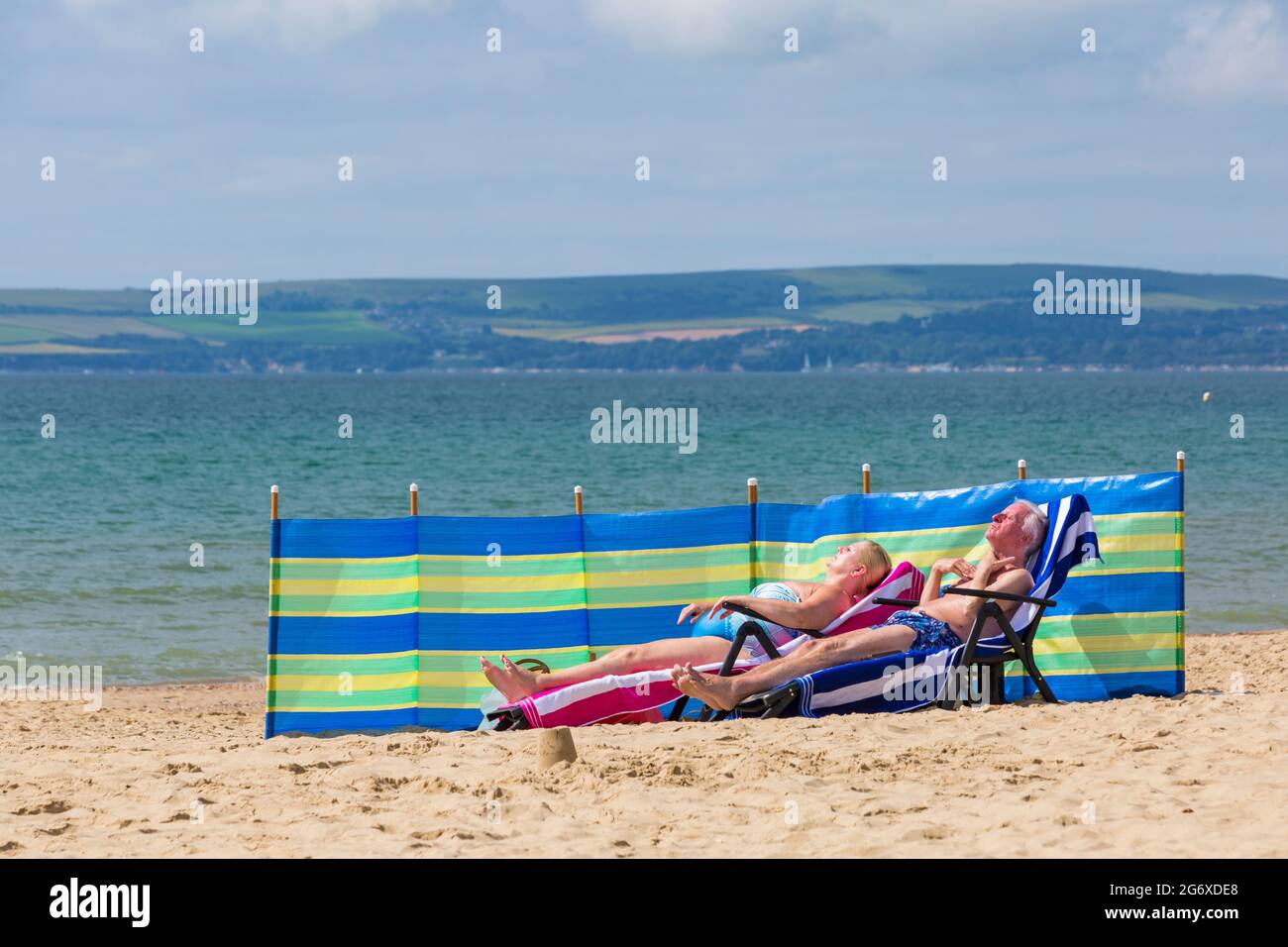 Bournemouth, Dorset UK. 9th July 2021. UK weather: lovely warm sunny, but humid day at Bournemouth beaches, as sunseekers head to the seaside to enjoy the sunshine. Mature couple sunbathing on sunloungers  with windbreak wind break windbreaker on sand on beach. Credit: Carolyn Jenkins/Alamy Live News Stock Photo