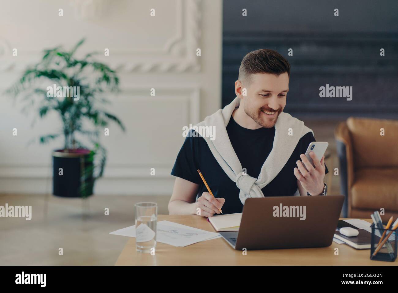 Happy bearded young man studies indoor with modern gadgets writes down notes from internet website makes records in notebook holds smartphone poses at Stock Photo