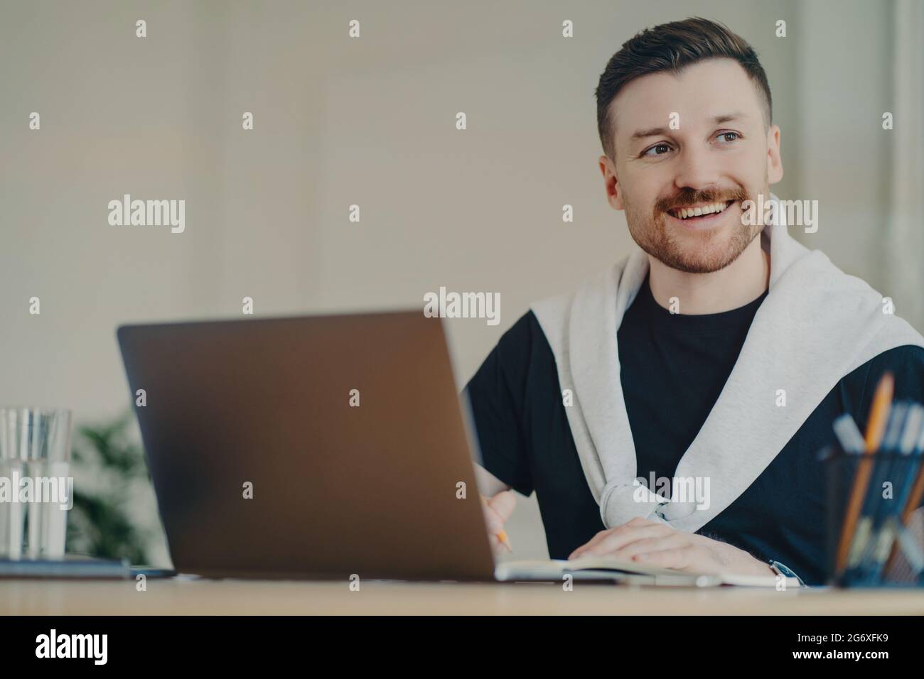 Happy bearded man office worker writes organisation plan works remotely makes notes of info sits in front of opened laptop looks away dressed in Stock Photo