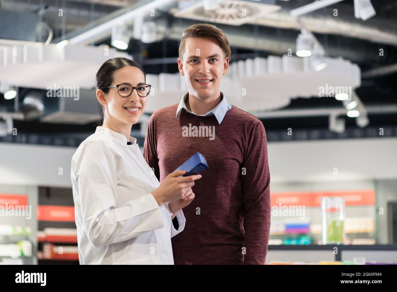 Portrait of handsome young man smiling as a happy customer, while standing next to a female reliable and friendly pharmacist in the interior of a cont Stock Photo