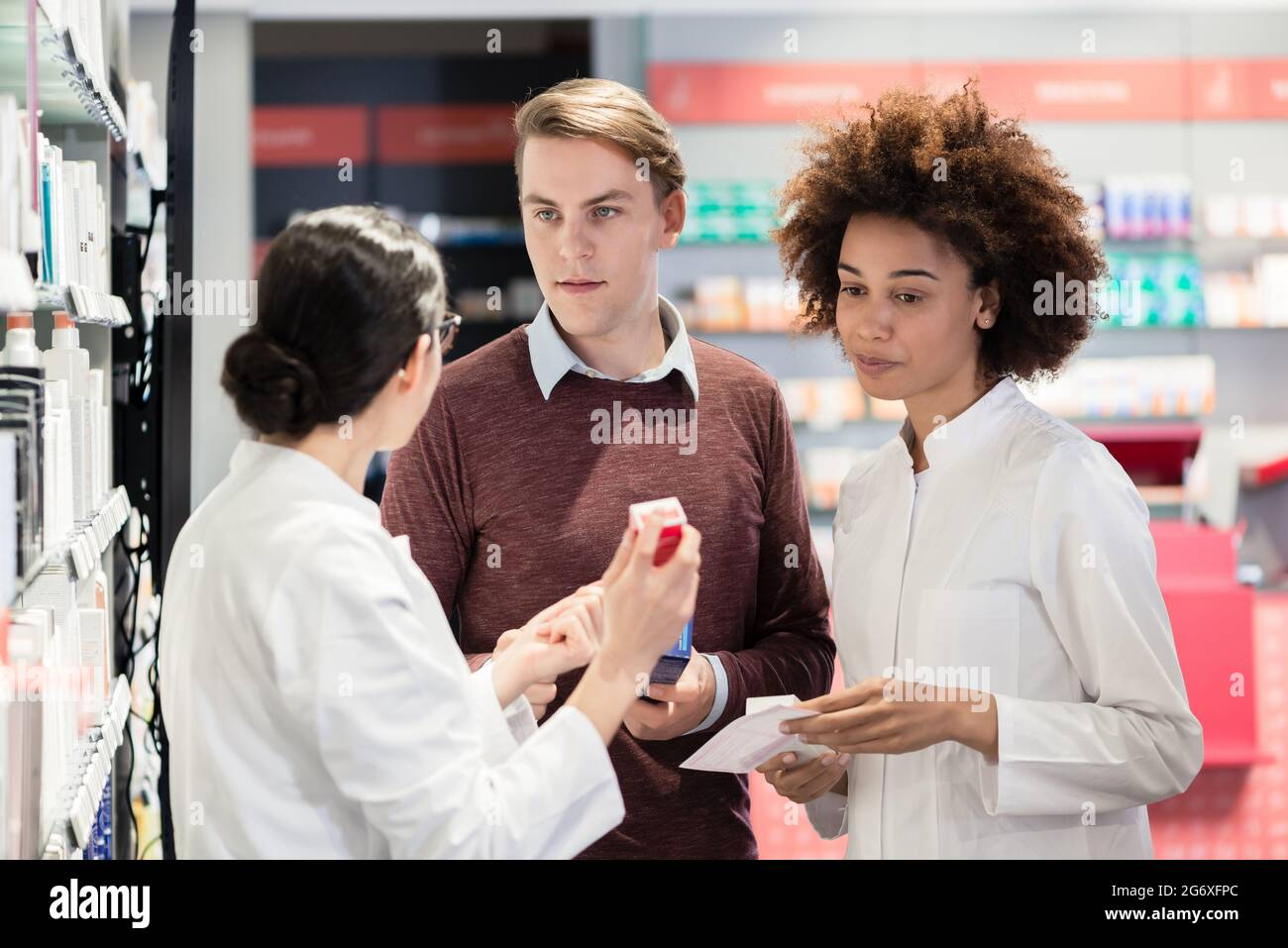Young man asking for advice and opinion from two experienced female pharmacists regarding a prescribed pharmaceutical product in the interior of a mod Stock Photo