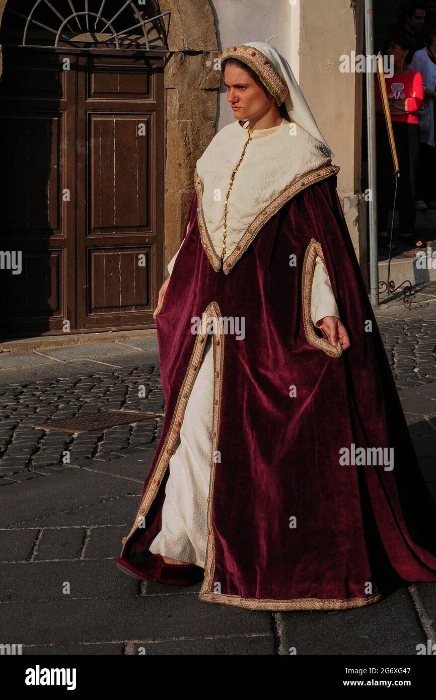 A young woman wearing the costume of a 14th or 15th century Italian lady or  noblewoman in a velvet overgown or surcoat trimmed with jewelled brocade,  stares resolutely ahead as she walks