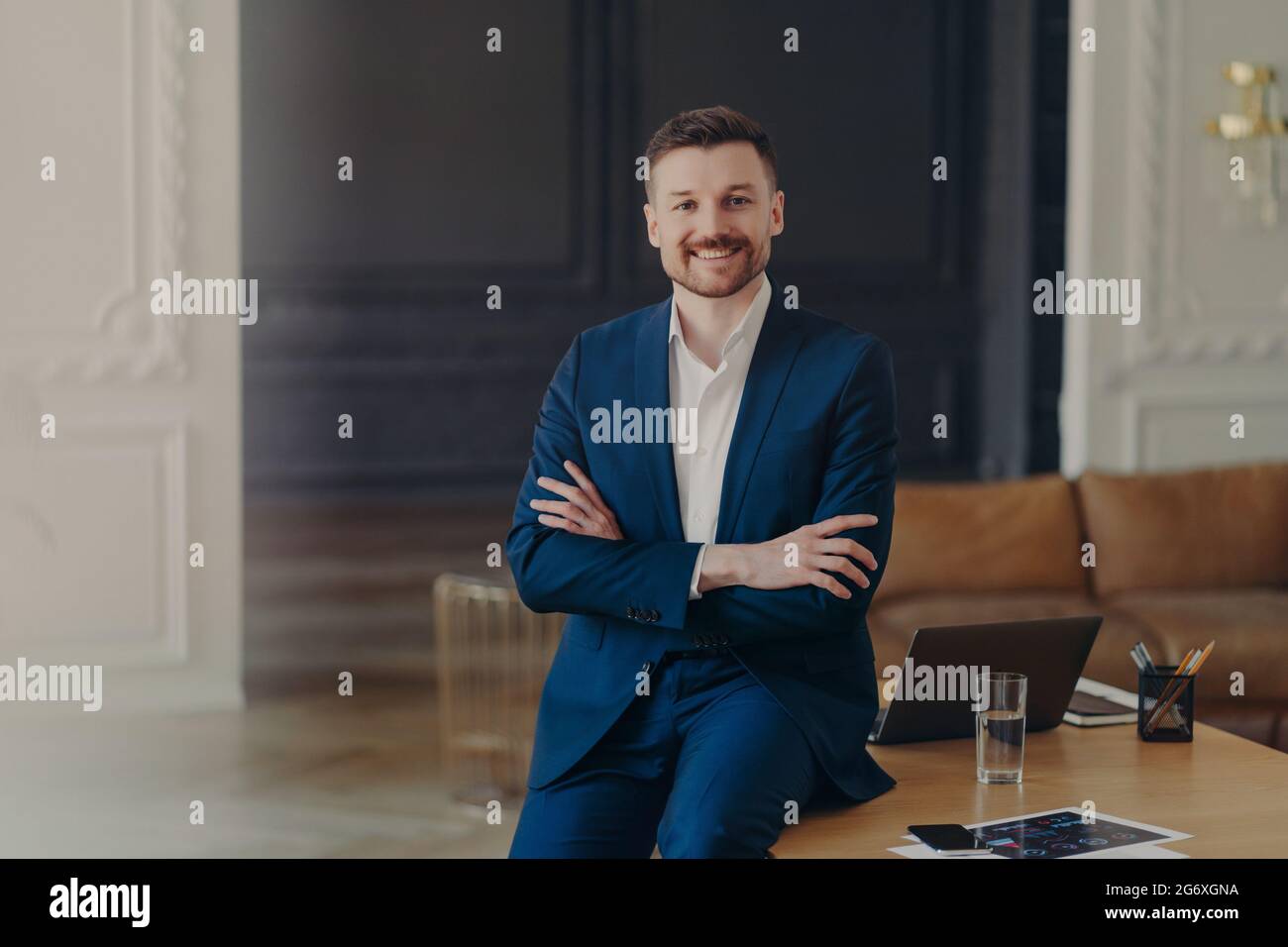 Photo of satisfied bearded prosperous businessman stands self assured in own cabinet or contemporary office keeps arms folded dressed formally smiles Stock Photo