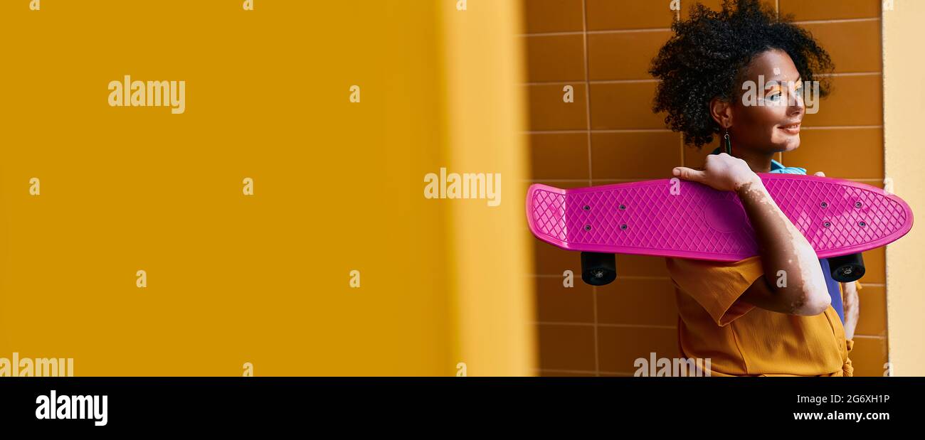 Youth culture. African American woman with purple penny board in hands near city building. Yellow urban wall with empty space for text Stock Photo