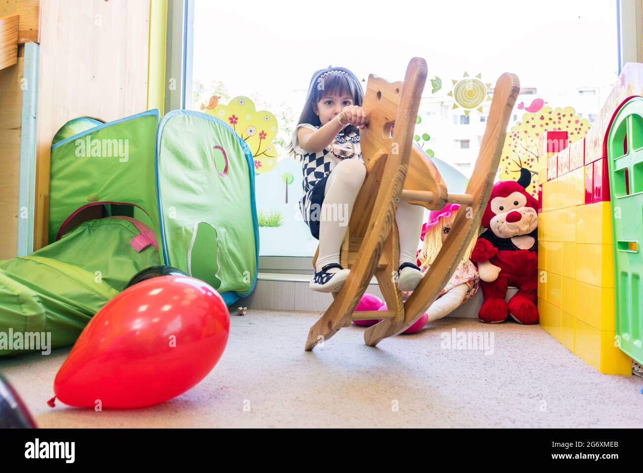 Cute pre-school girl looking at camera while swinging on a vintage wooden horse during free playtime at the kindergarten Stock Photo