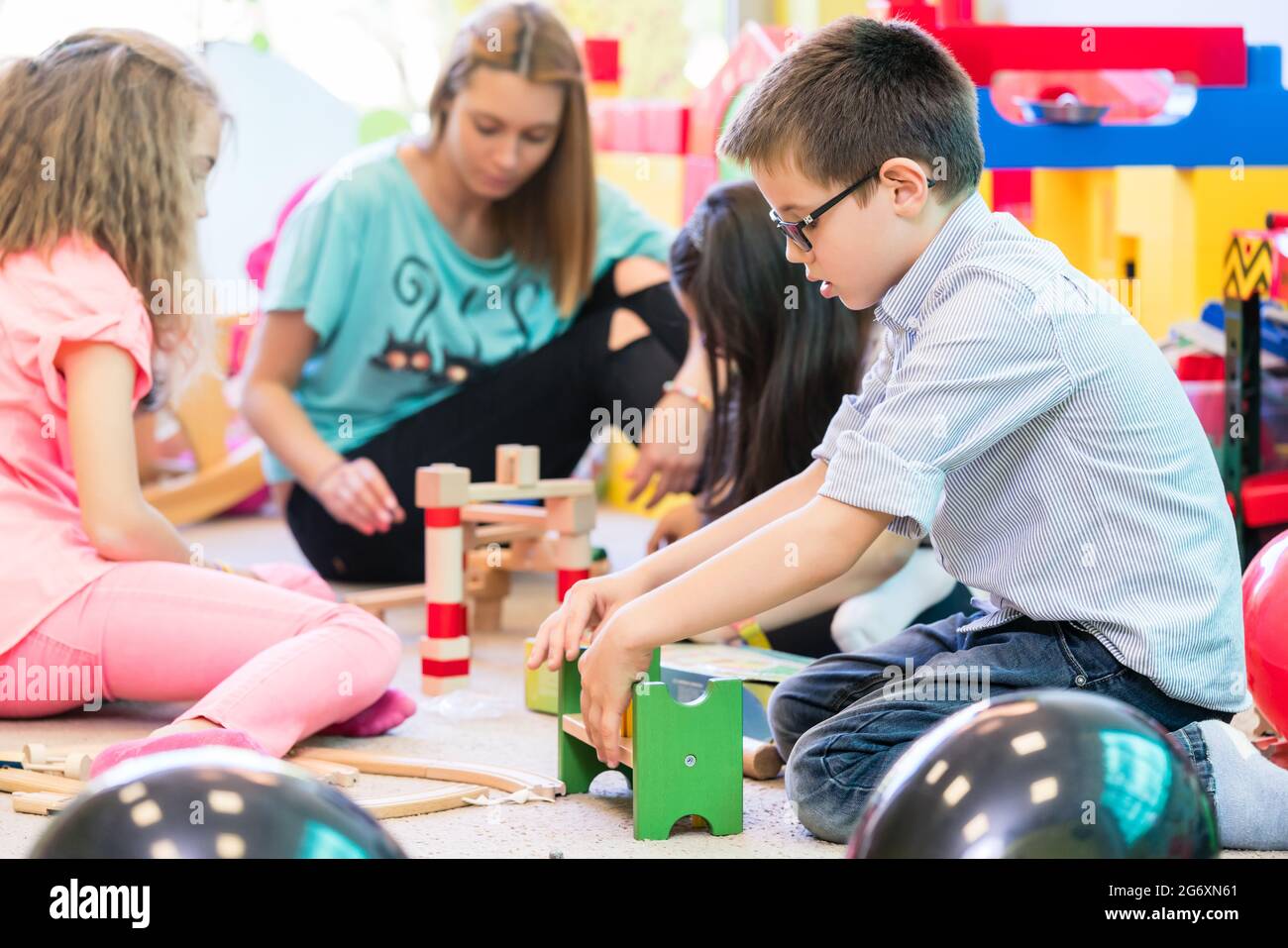 Cute pre-school boy cooperating with his colleagues at the construction of a structure made of wooden toy, blocks under the guidance of a young kinder Stock Photo