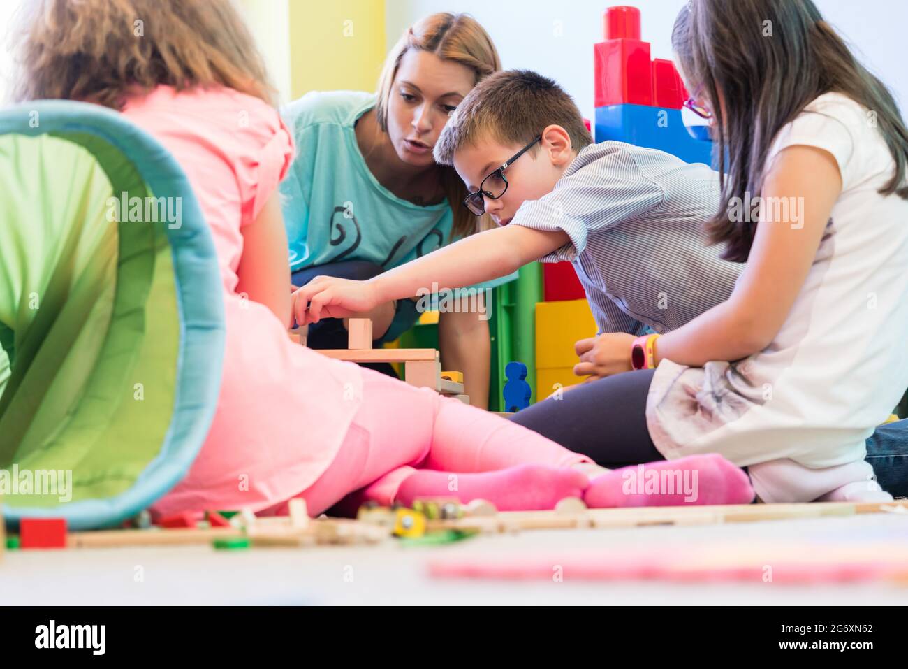 Cute pre-school boy cooperating with his colleagues at the construction of a structure, made of wooden toy blocks under the guidance of a young kinder Stock Photo