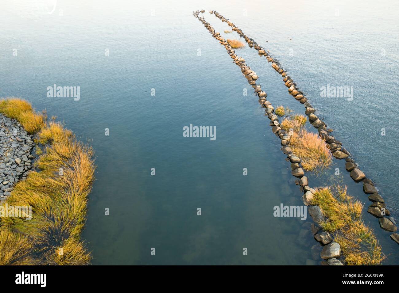 Aerial of calm water with a row of large rocks covered in sea grass jutting out into the bay. Stock Photo