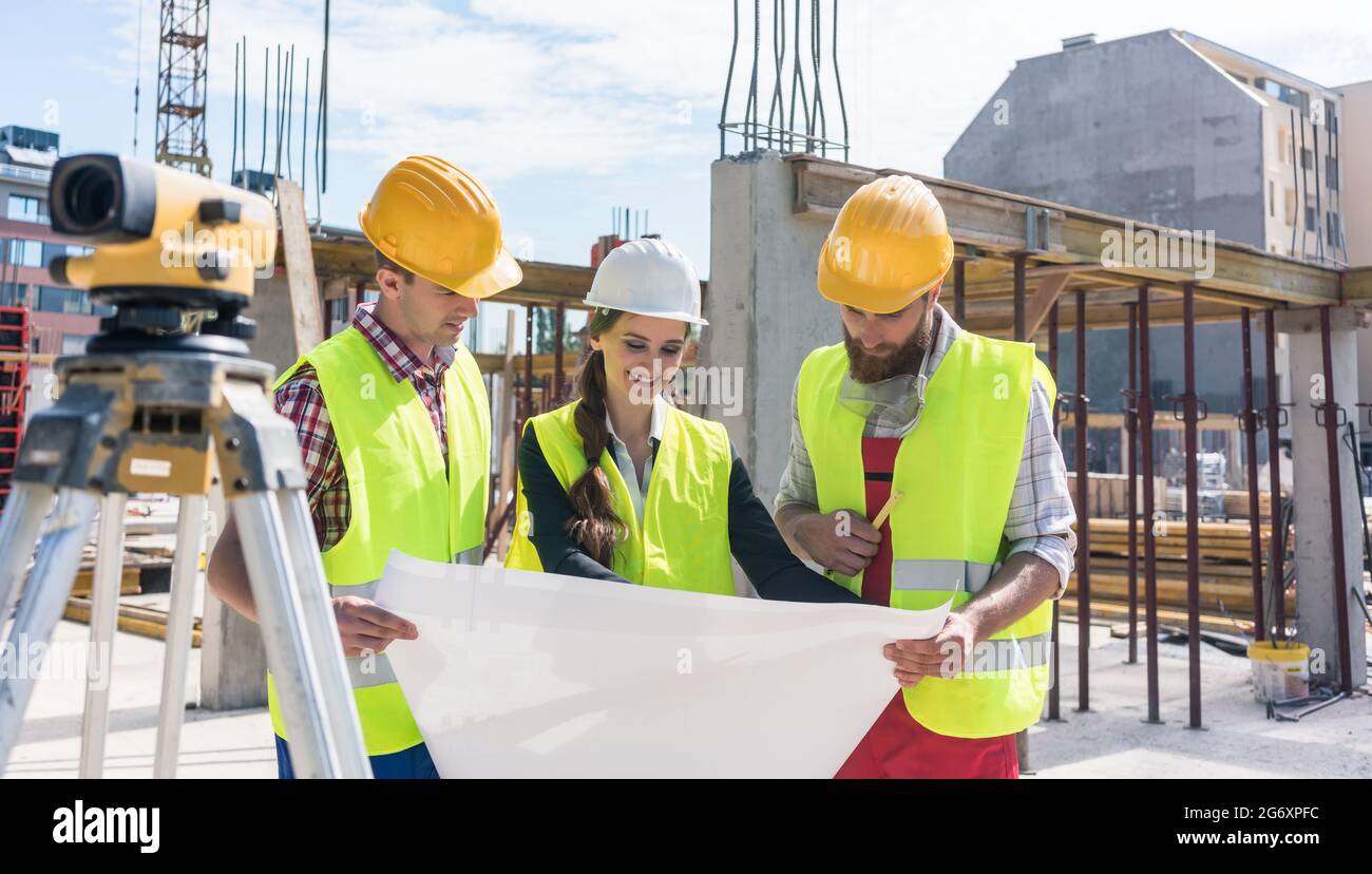 Experienced female architect smiling while analyzing a blueprint together with two workers outdoors on the construction site of a modern residential b Stock Photo