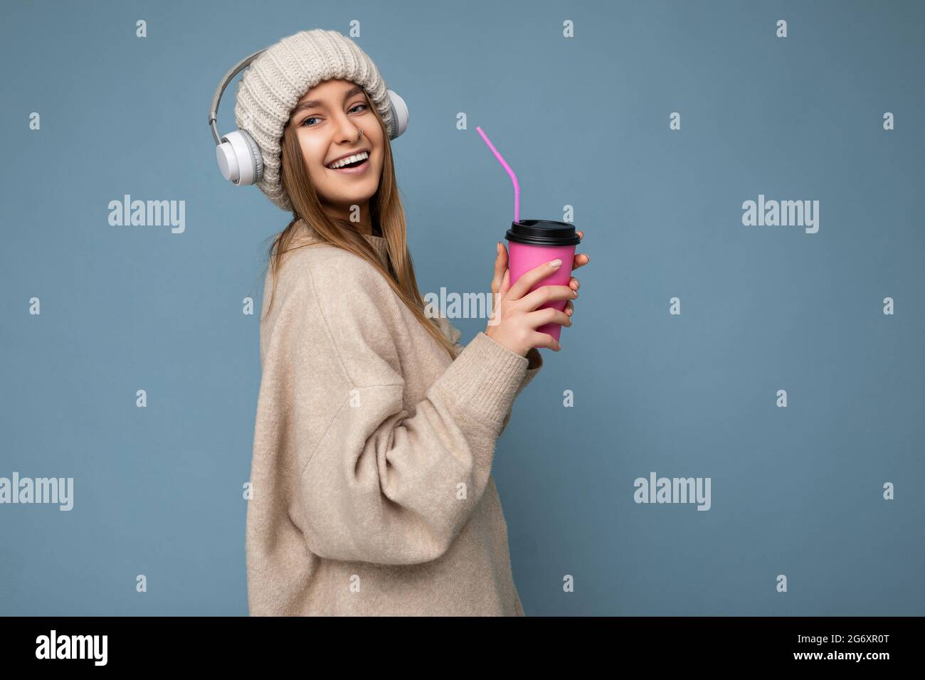 Beautiful happy smiling young blonde female person wearing beige winter sweater and hat isolated over blue background drinking coffee wearing white Stock Photo