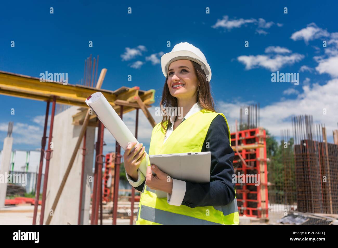 Portrait of a confident female architect or engineer with can-do attitude smiling while holding a rolled blueprint and a tablet on the construction si Stock Photo