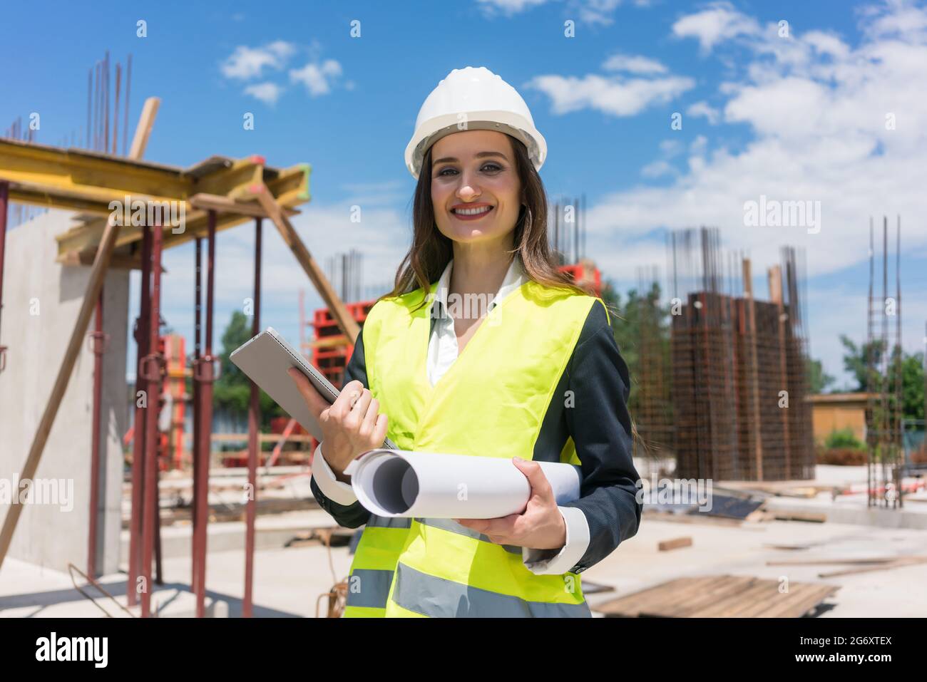 Portrait of a confident female architect or engineer with can-do attitude smiling while holding a rolled blueprint and a tablet on the construction si Stock Photo