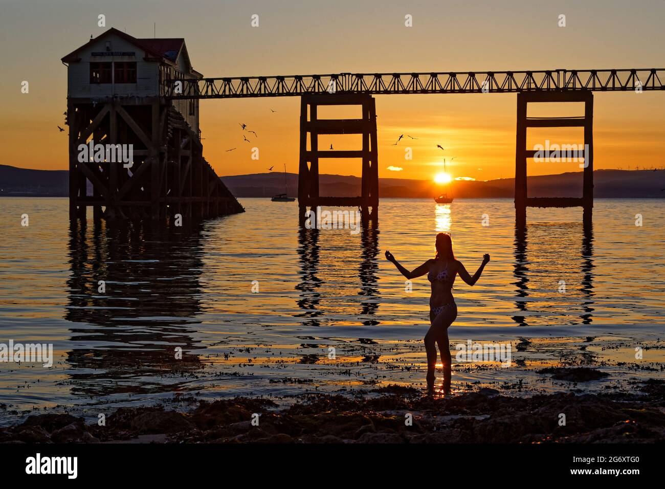 Pictured: Natasha Jenkins in the sea during sunrise as seen through the RNLI boat station in Mumbles, near Swansea, Wales, UK. Sunday 13 June 2021 Re: Stock Photo