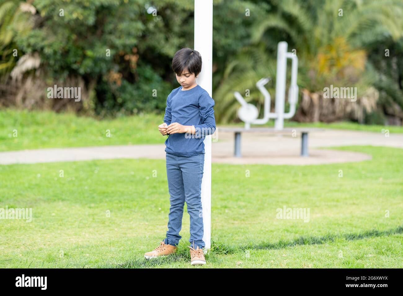 Sad kid standing alone looking down at park. Lonely young boy holding shred of grass in his hands. Stock Photo