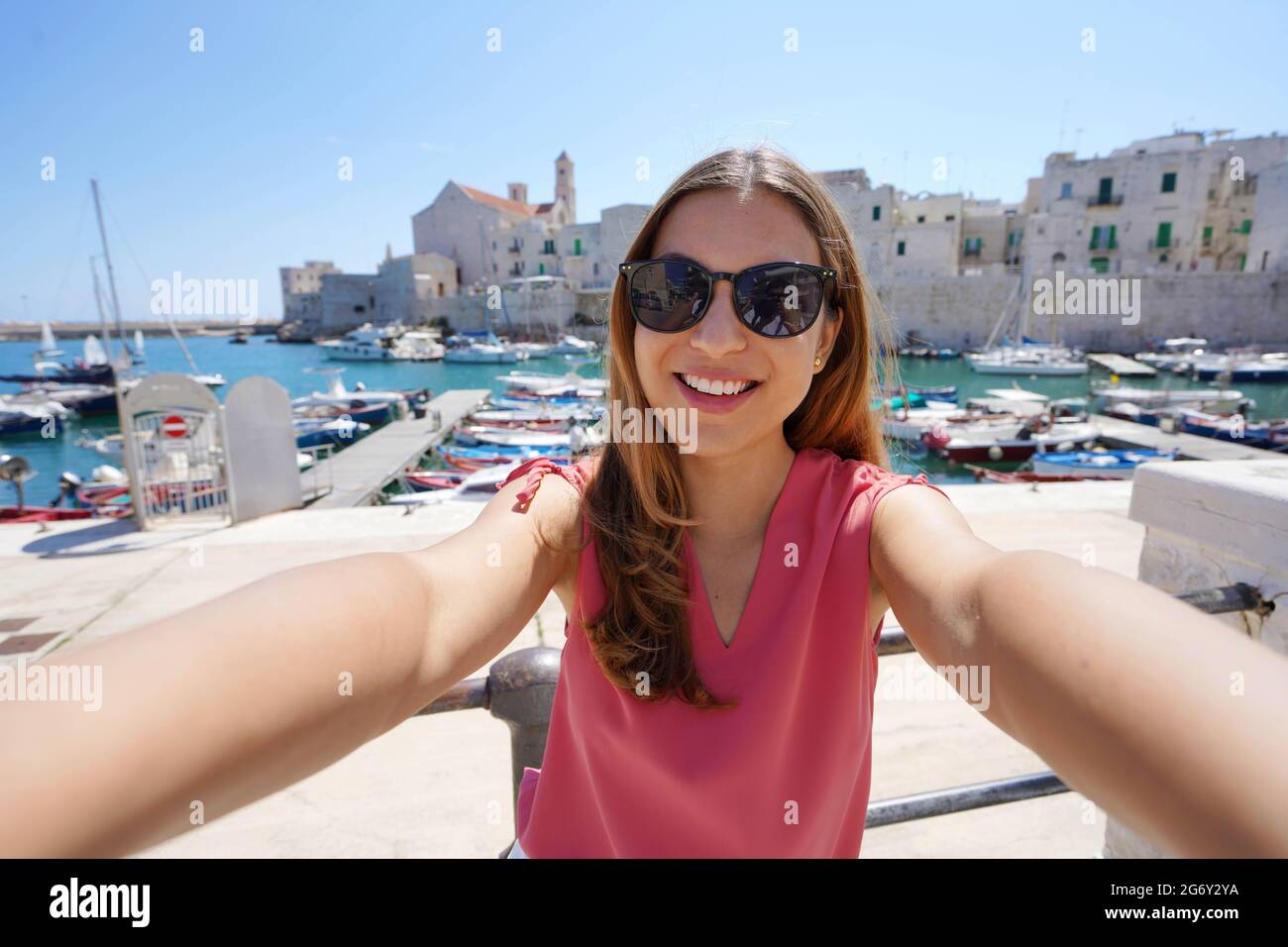 Happy beautiful woman with sunglasses taking selfie smiling at the camera with the old town of Giovinazzo on the background in southern Italy Stock Photo