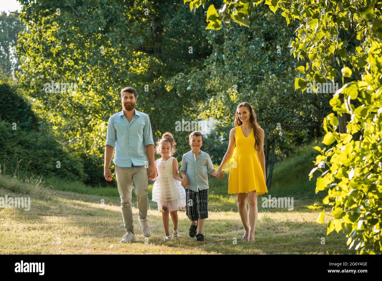 Full length view of a happy family with two children wearing casual summer clothes while holding hands during recreational walk in the park Stock Photo