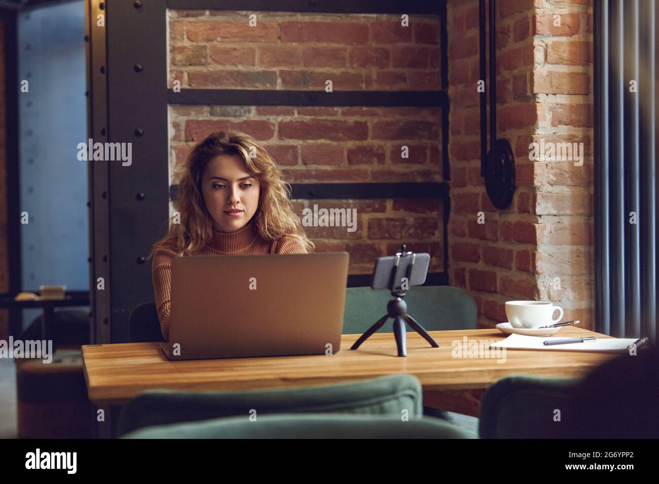 Woman working on laptop at table with smartphone on tripod Stock Photo