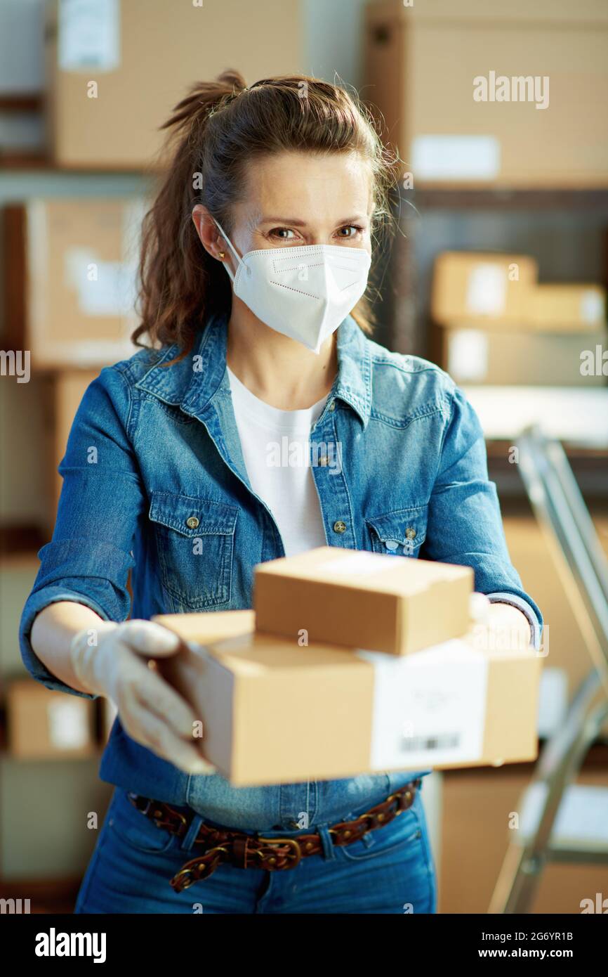 Delivery business. modern female in jeans with ffp2 mask giving parcel in the warehouse. Stock Photo
