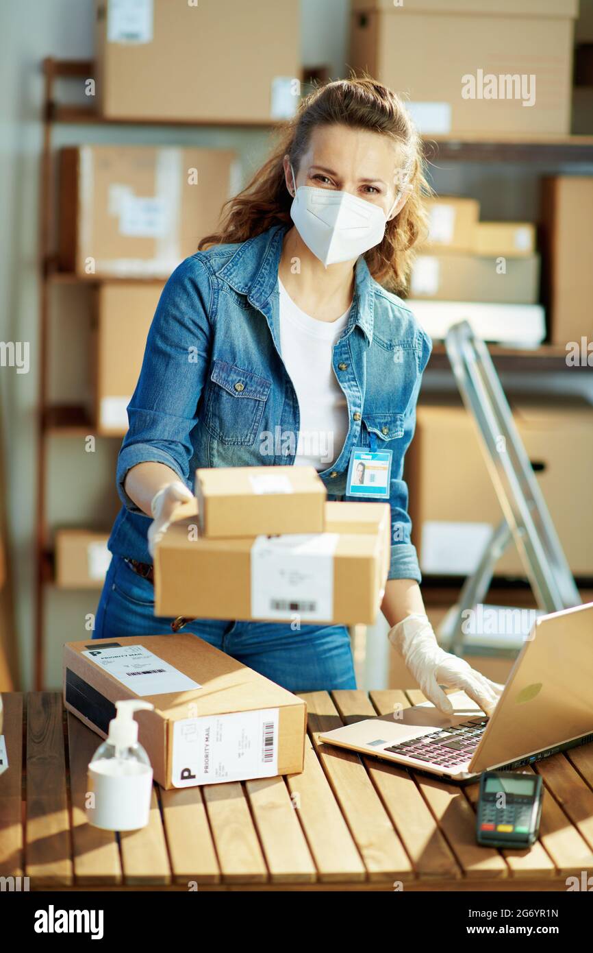 Delivery business. modern woman in jeans with laptop and ffp2 mask giving parcel in the office. Stock Photo