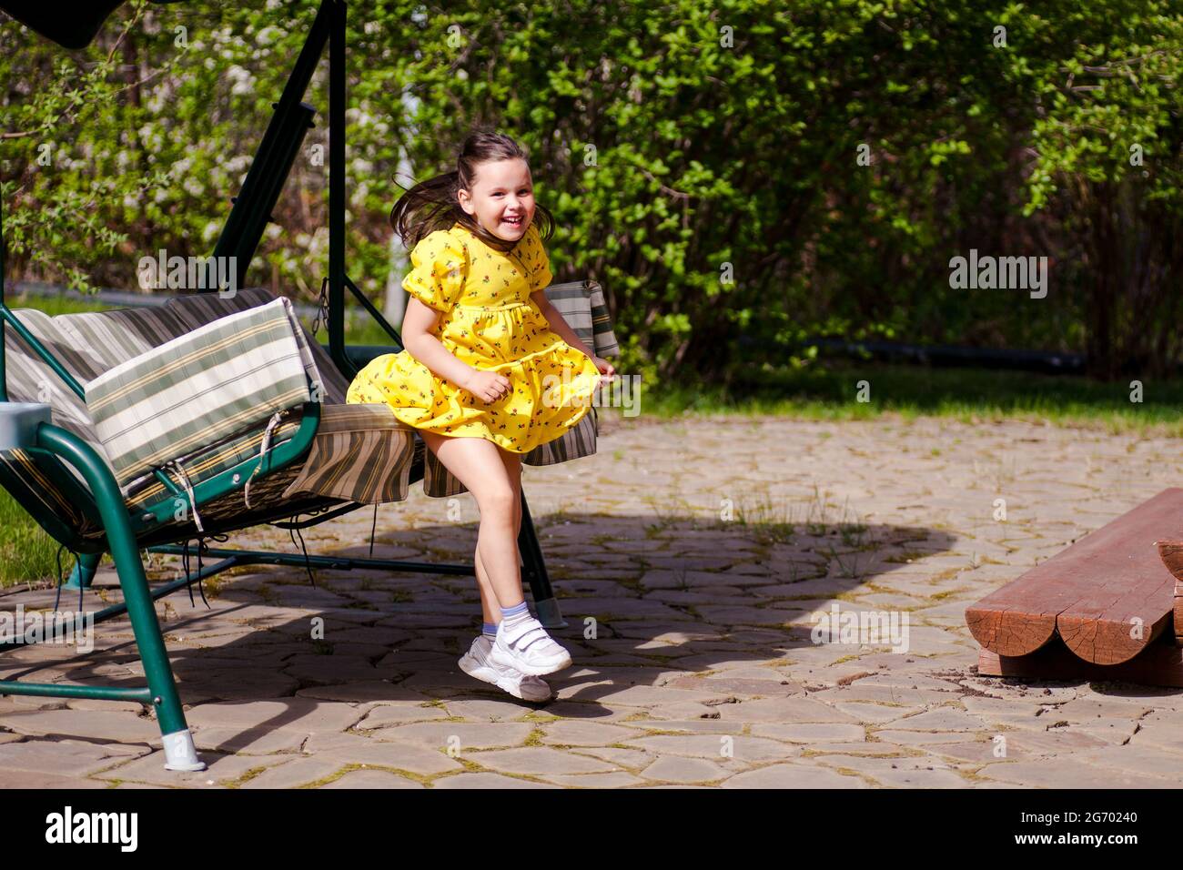 frozen moment of movement or jump, a smiling girl in a yellow dress jumps into the air from a garden swing on a sunny day on a family vacation Stock Photo