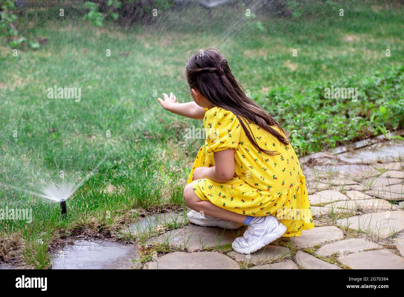 rear view of a girl who is playing with water drops from a lawn sprinkler, a girl helps her parents to water the grass on the lawn Stock Photo